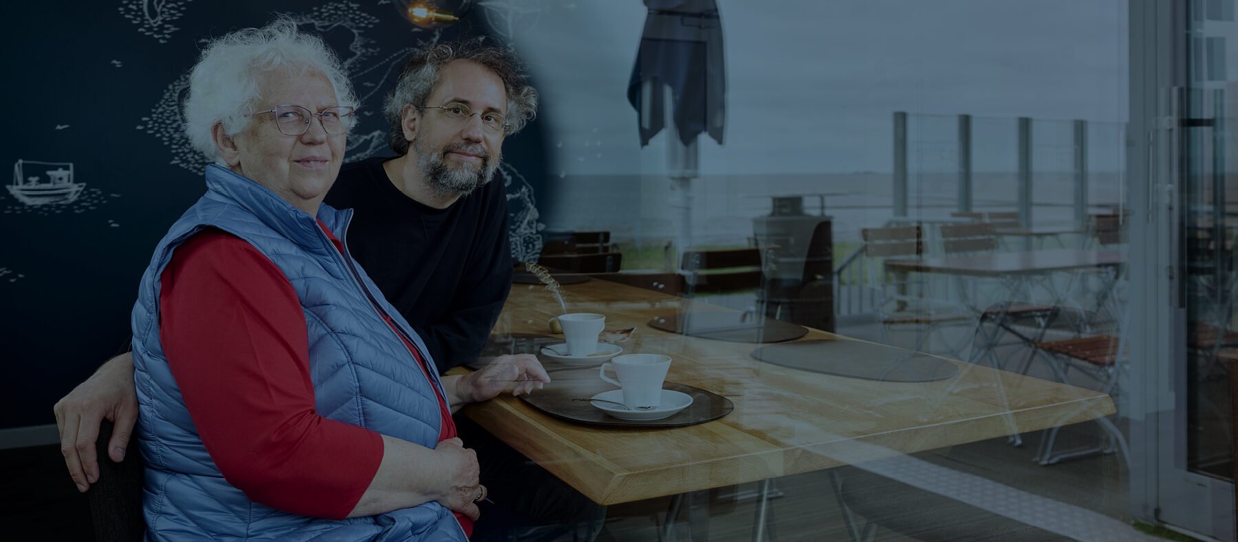 Zwei Personen sitzen an einem Tisch in einem Café mit Blick auf das Meer durch ein Fenster.