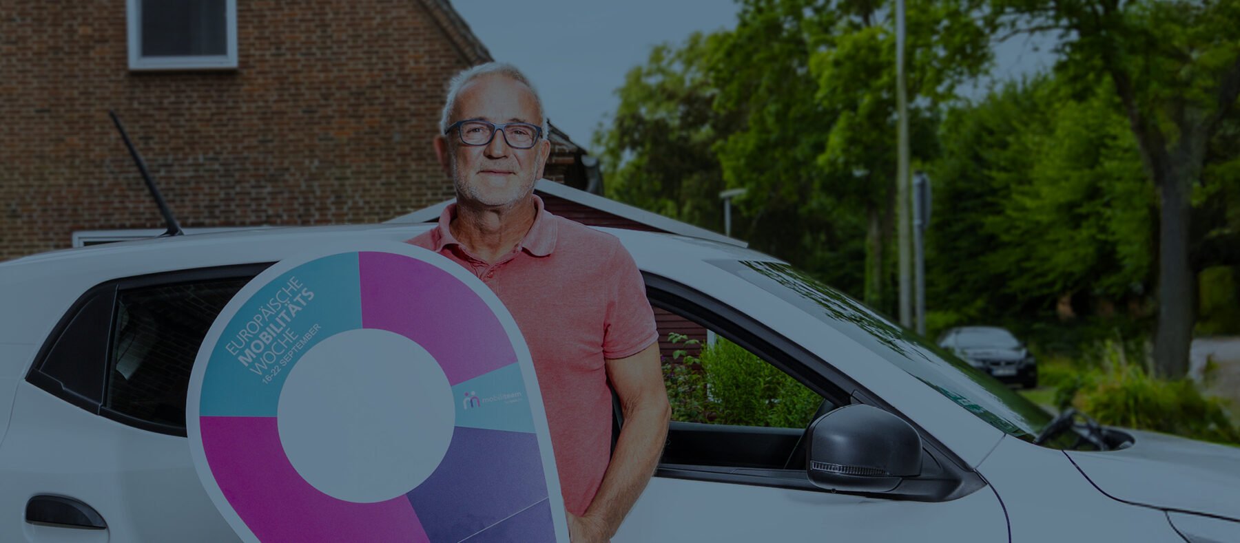 Uwe Eisenmann steht vor einem weißen Auto mit der Aufschrift „Dörpsmobil SH Friedrichstadt“ und hält ein großes, buntes Schild.
