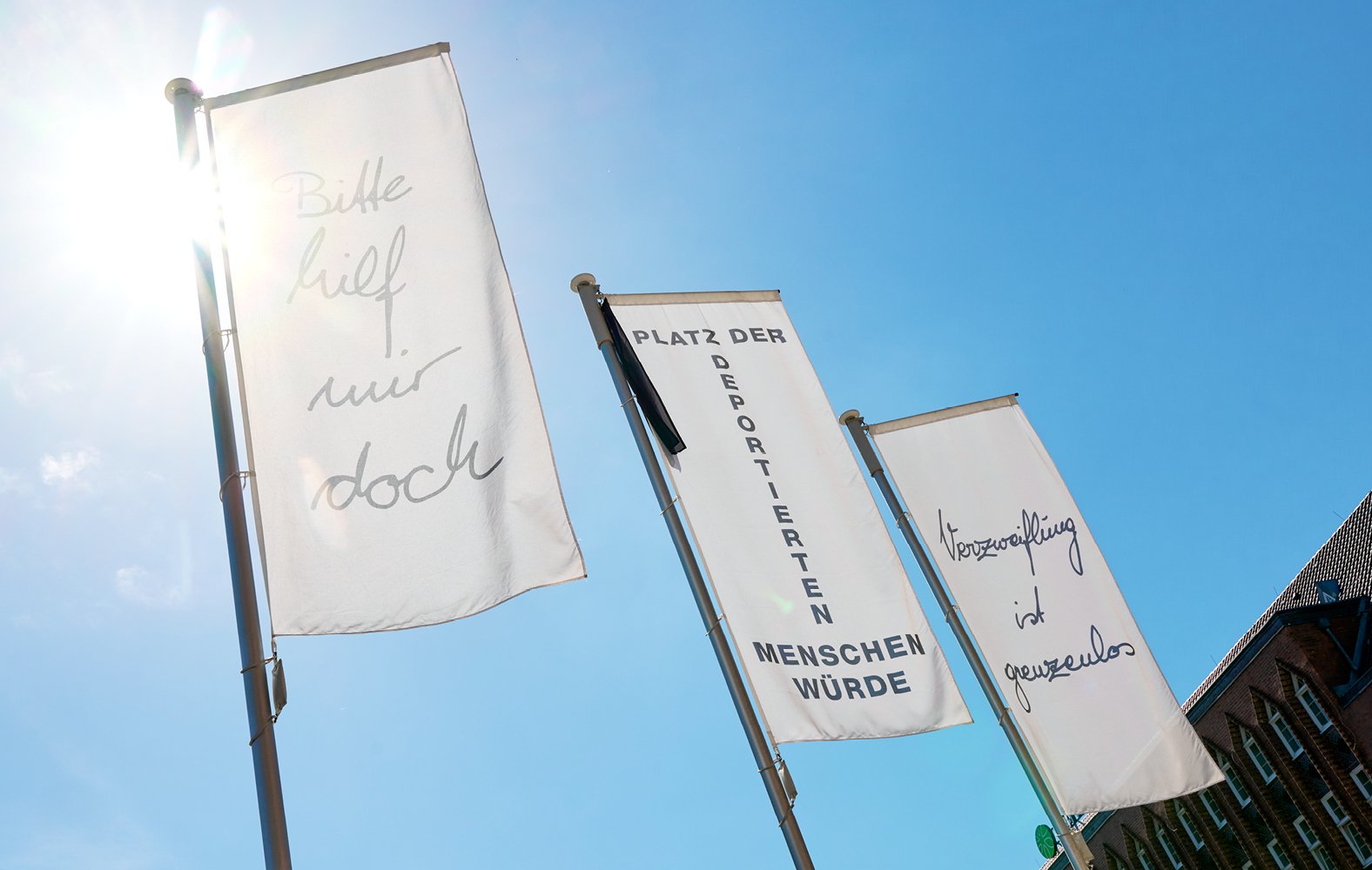 Weiße Fahnen mit Aufschriften wehen vor blauem Himmel. Ein Gebäude mit Giebeldach ist im Hintergrund zu sehen.