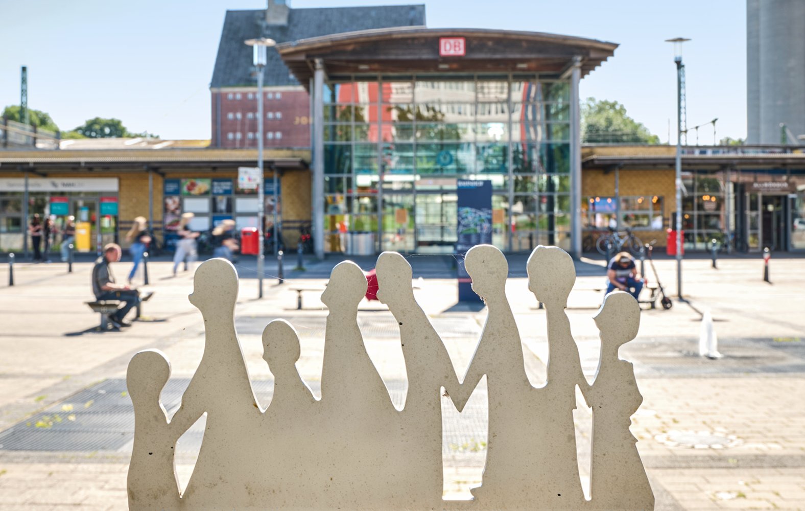 Skulptur aus Silhouetten von Menschen vor einem modernen Gebäude mit Glasfassade und roten Strukturen.