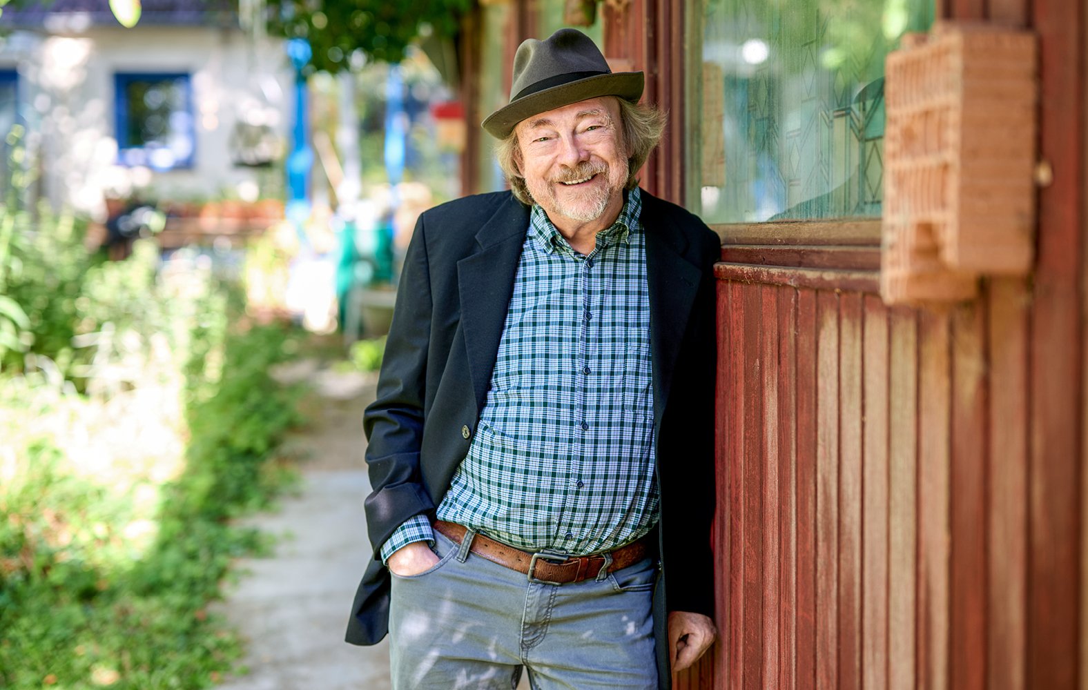Heino Schomaker mit Hut lehnt an einer Holzwand, trägt ein kariertes Hemd und lächelt. Im Hintergrund ist ein Weg mit Pflanzen zu sehen.