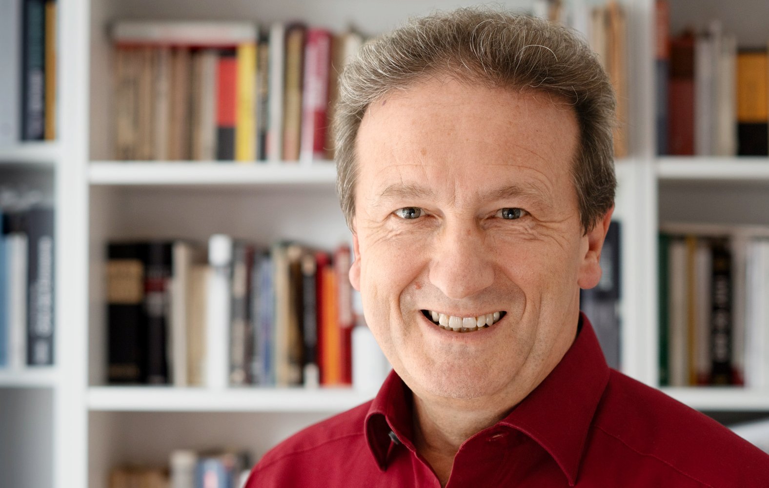 Dr. Harald Schmidt in rotem Hemd lächelt vor einem Bücherregal.
