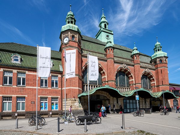 Fahrräder stehen auf gepflastertem Platz vor einem großen roten Backsteingebäude mit grüner Überdachung. Menschen gehen hinein und hinaus.