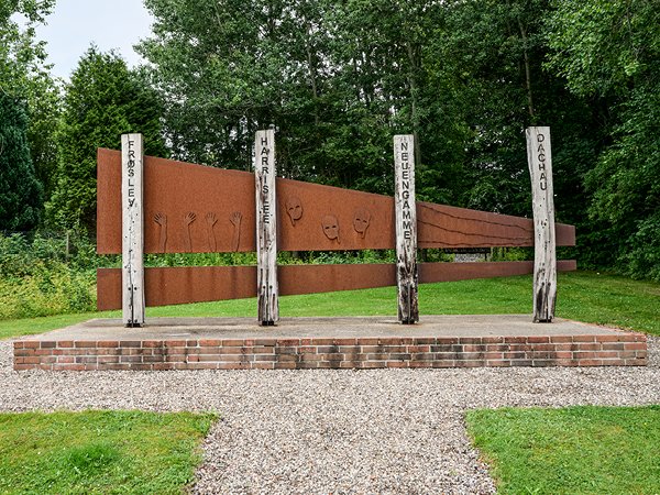 Denkmal mit vier vertikalen Holzstämmen auf einem Ziegelpodest, umgeben von Bäumen und Rasen, im Hintergrund eine rostfarbene Metallwand